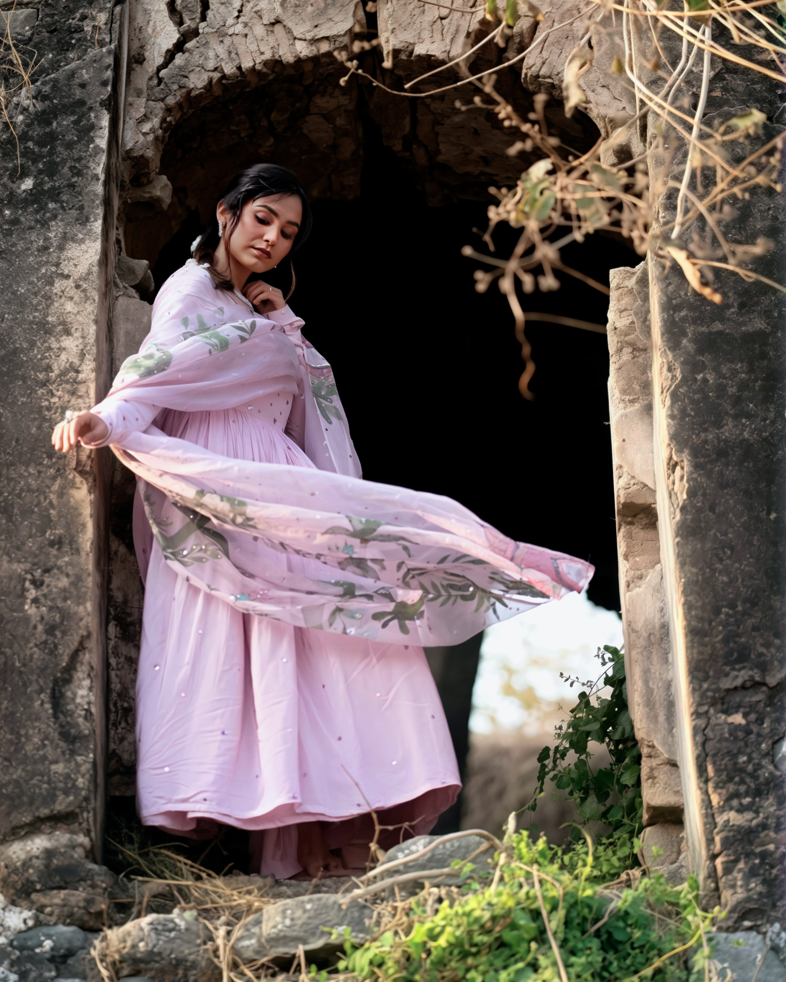 Woman in a pink dress with floral patterns standing in an old stone archway.